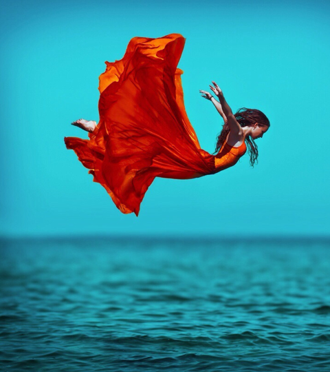 Woman in red flowy dress floating above water with her arms outstretched like she is flying representing celebrating the Year that was 2022 by taking a leap of faith.  