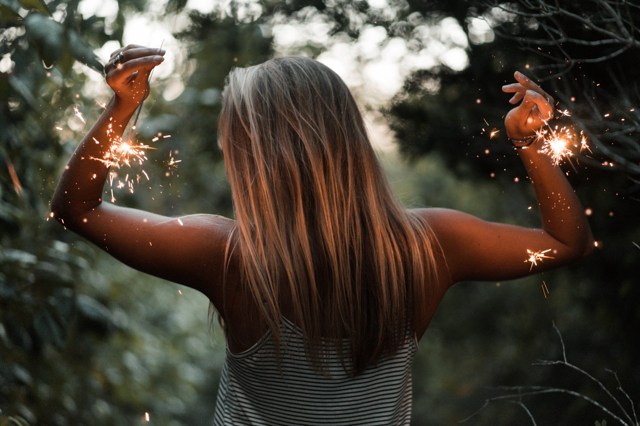 A woman with long hair standing with her back to you in a forest clearing holding a sparkler in each hand