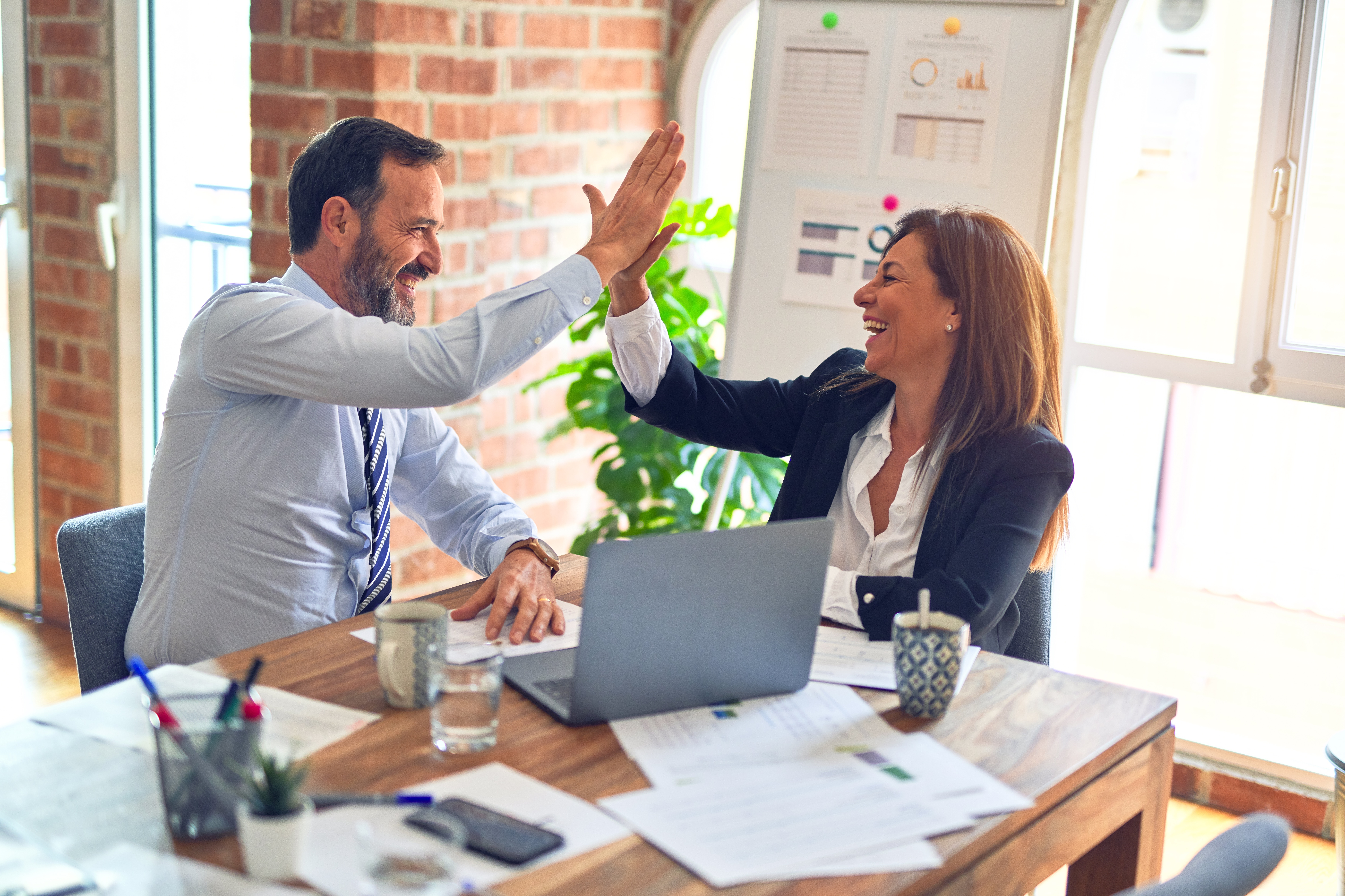 A man and a woman sitting at a table in front of a laptop high fiving to depict discovering the The Top 6 Most Impactful Habits for leaders.
