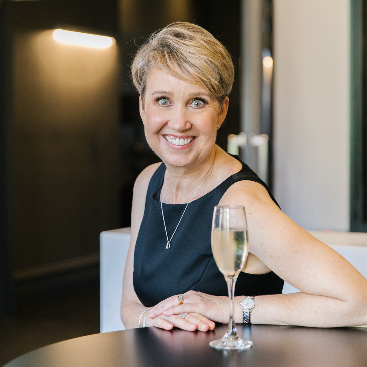 A photo of Sinja seated at a table with a glass of champagne looking at the camera.